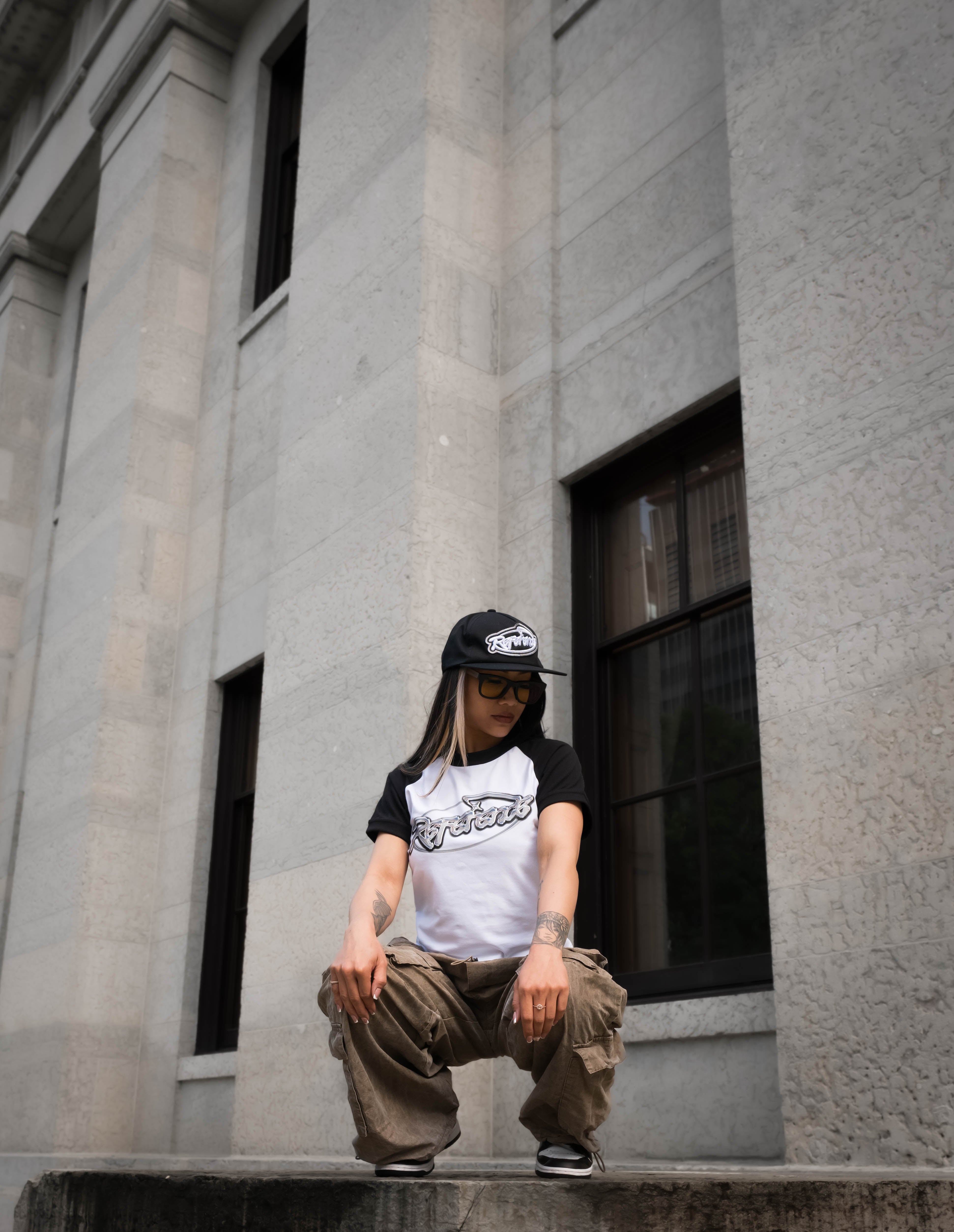 Person wearing a cap and sunglasses sitting on a building ledge.